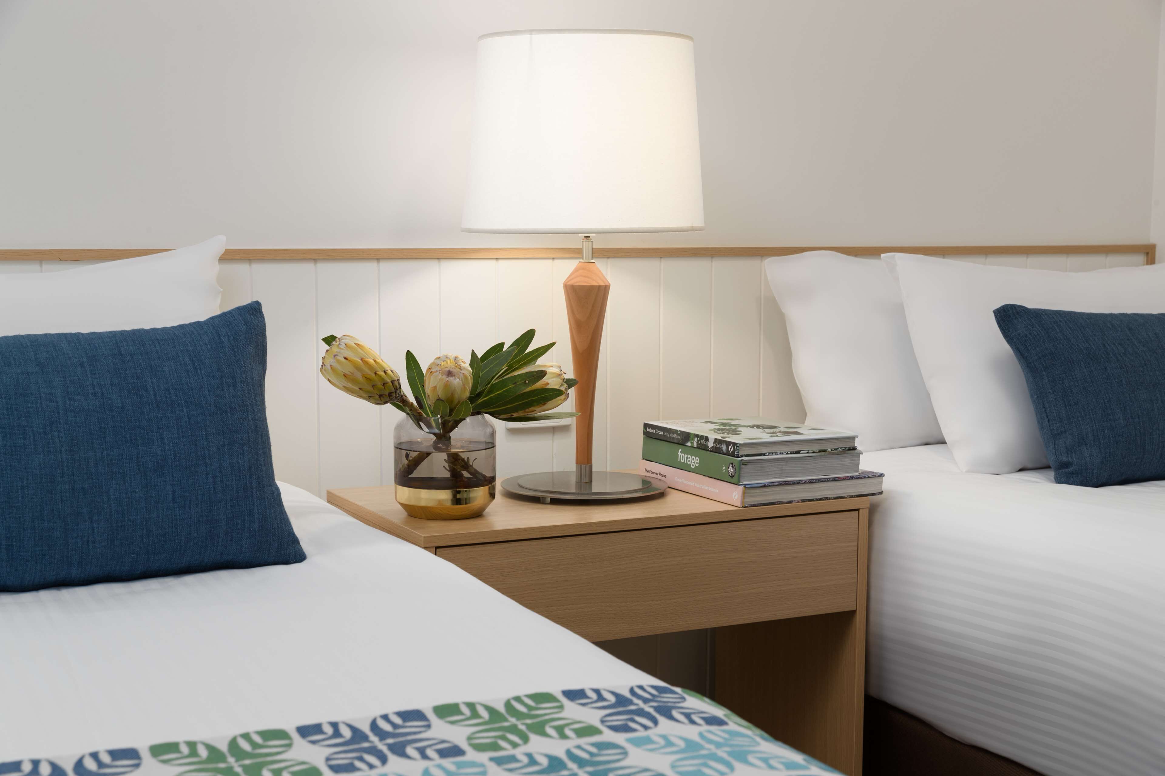 A hotel room with two beds, a bedside table with a lamp, a vase with flowers, and a stack of books.