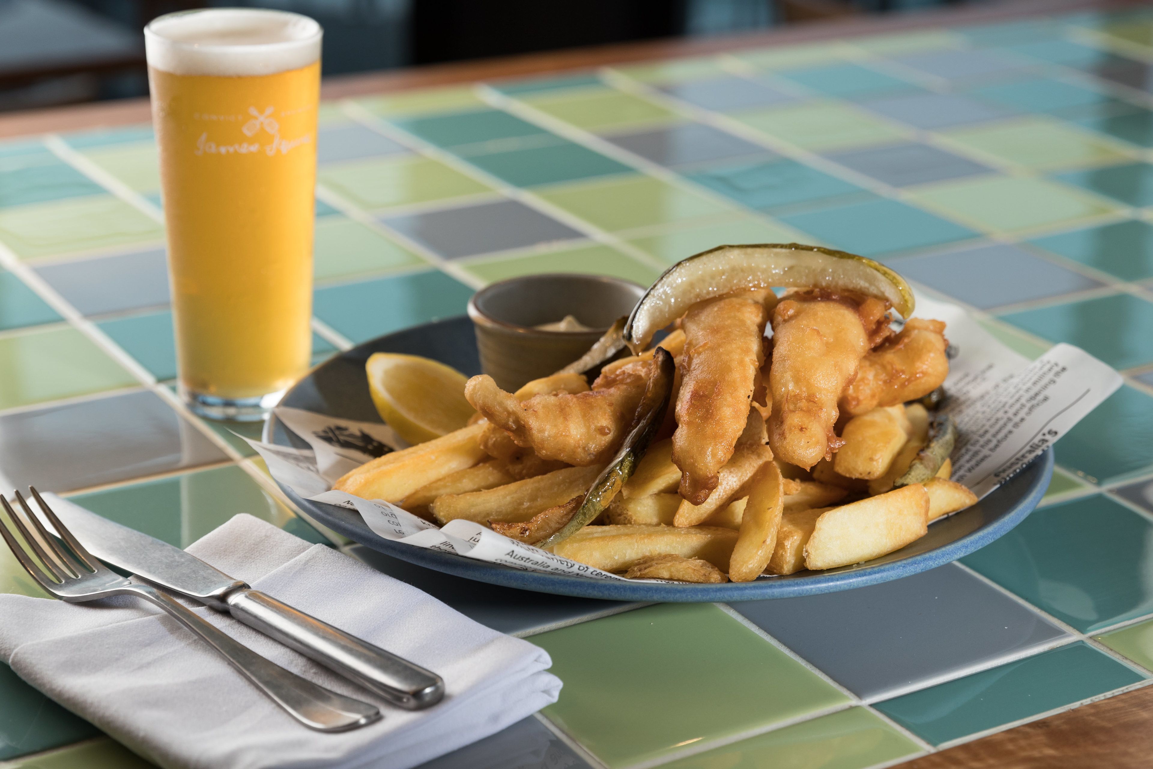 A plate of fish and chips with a side of tartar sauce and a glass of beer on a checkered table.