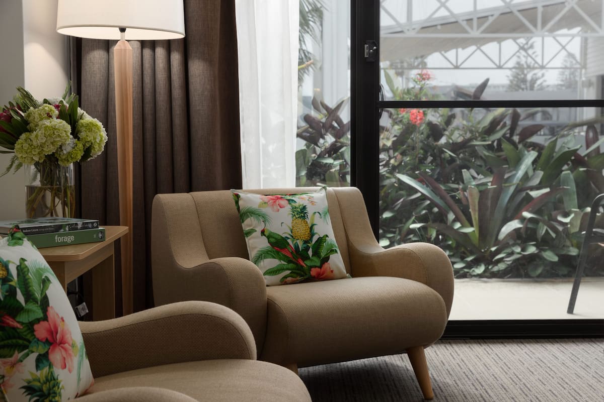 Une chambre d’hôtel avec un fauteuil beige, un coussin à motifs floraux, une table d’appoint avec une lampe, et une vue sur un balcon agrémenté de plantes.