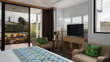 A hotel room with a large bed, a desk with a TV, two armchairs, a balcony with a view of the garden, and a large window with blinds.
