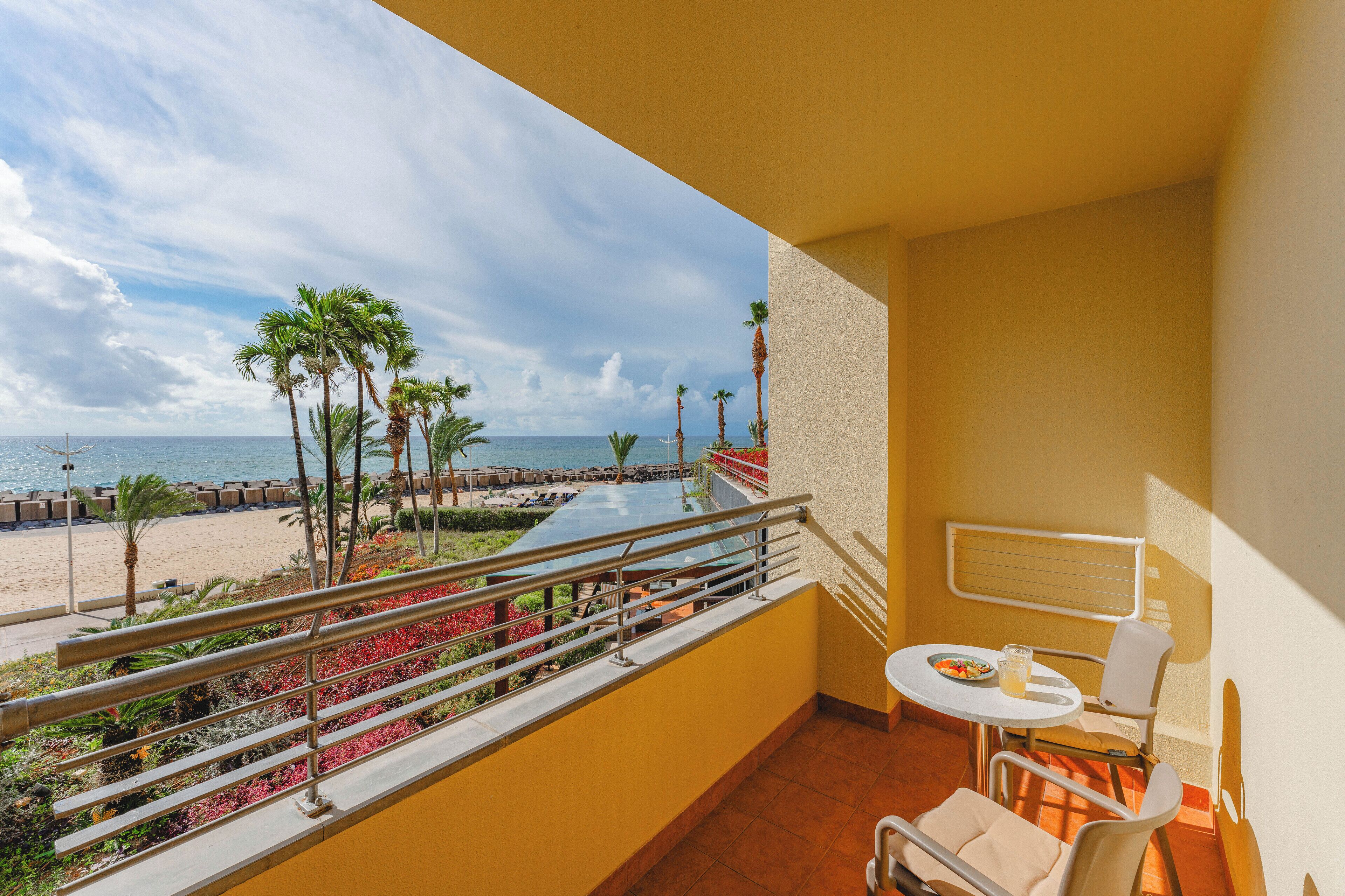 Uma varanda com vista para a praia, uma mesa com cadeiras e um corrimão.