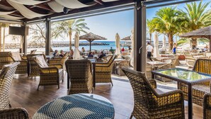 An outdoor seating area with wicker furniture and umbrellas, overlooking a beach and rocky shoreline. - Calheta Beach - All Inclusive (Calheta)