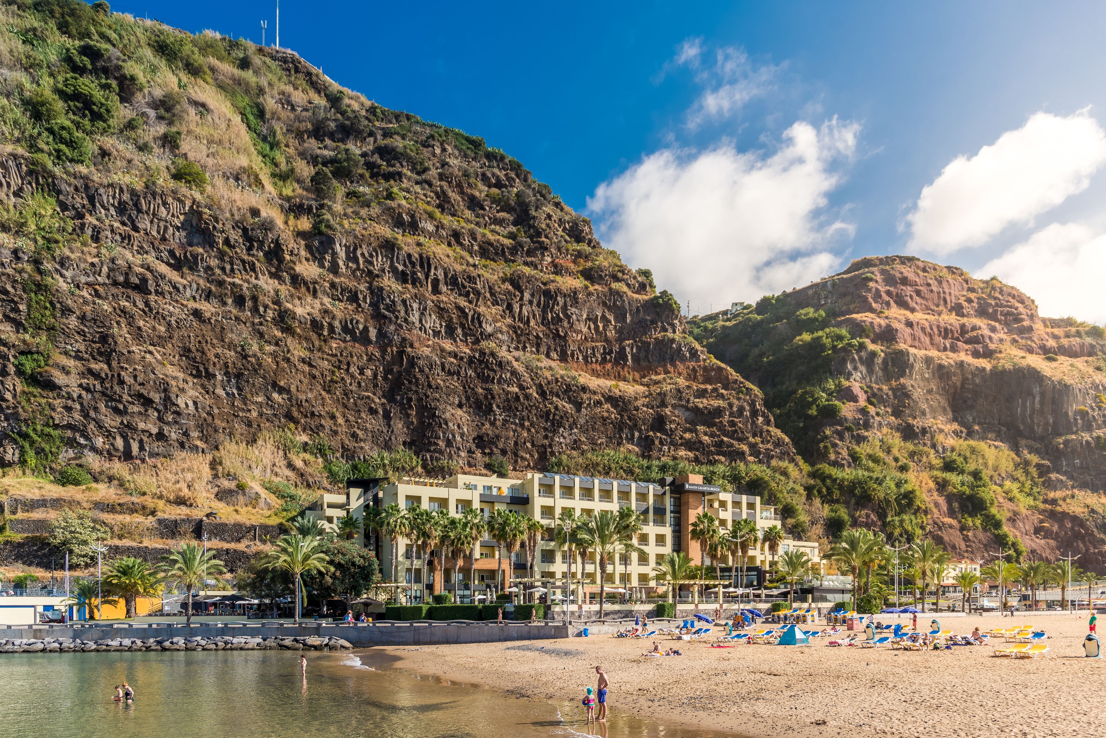 A coastal hotel with multiple floors, palm trees, and a beach in the foreground.