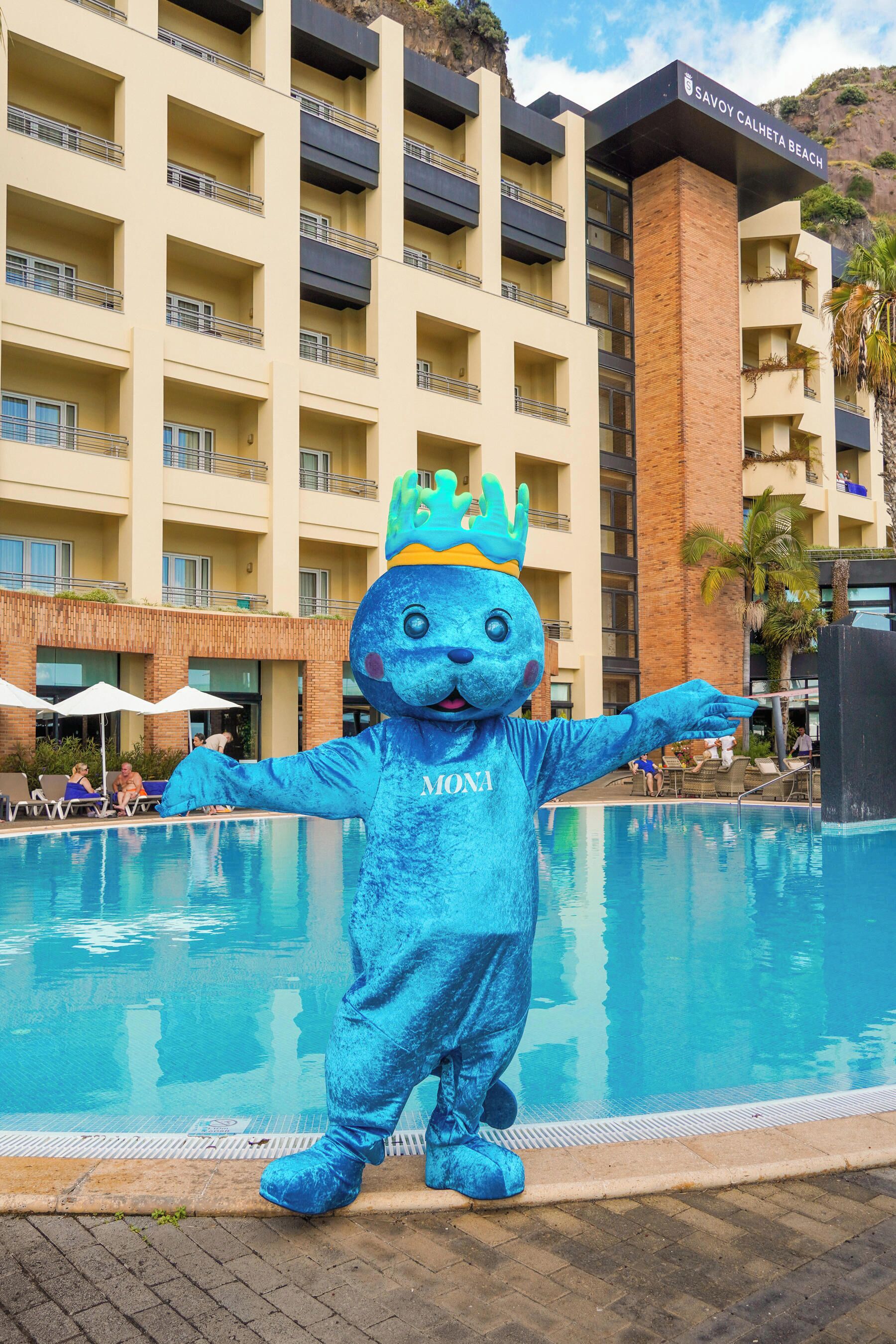 Un personaje con disfraz azul y corona de pie junto a una piscina, con un edificio de hotel al fondo.
