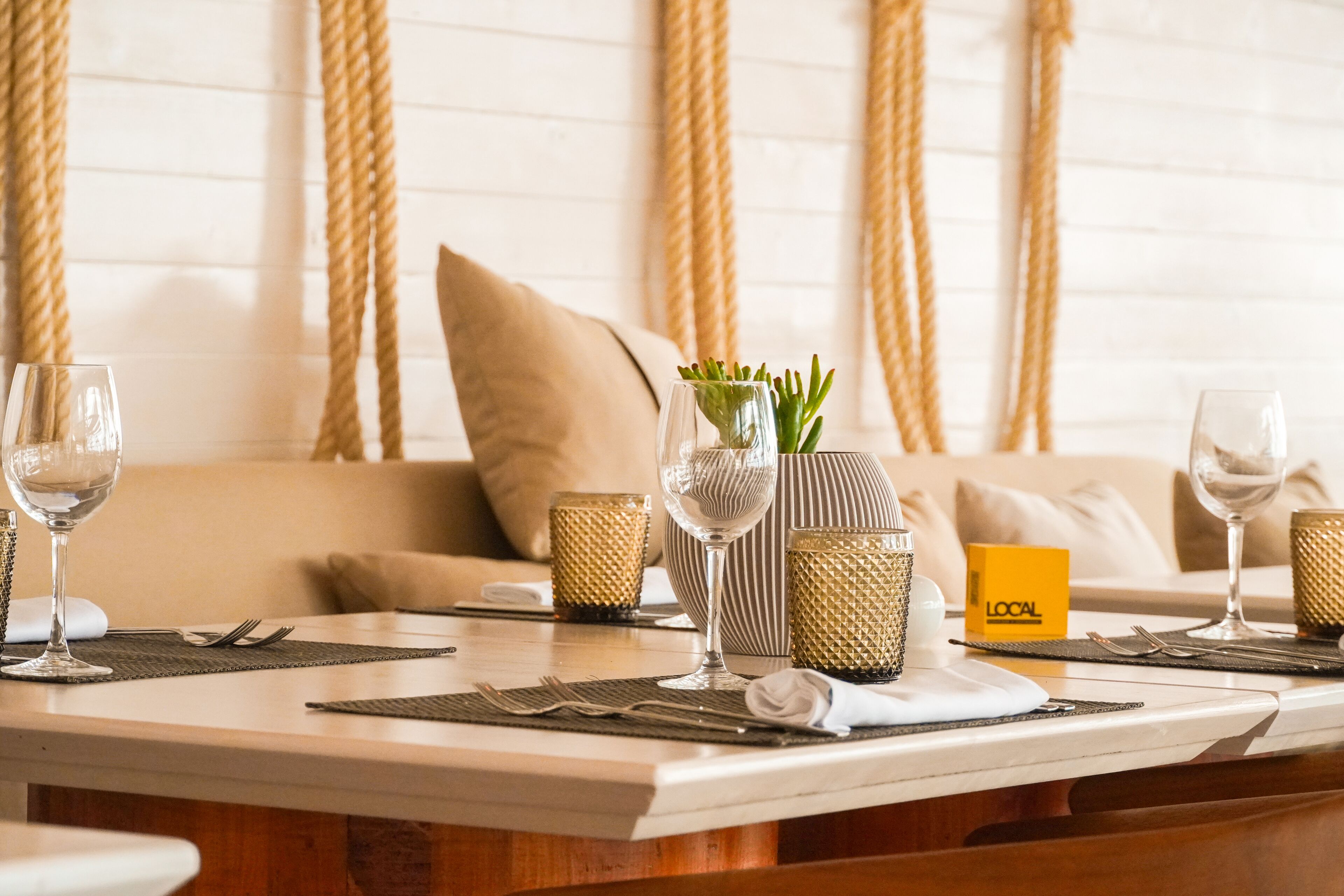 A dining table set with wine glasses, a vase with green plants, and a 'LOCAL' sign.
