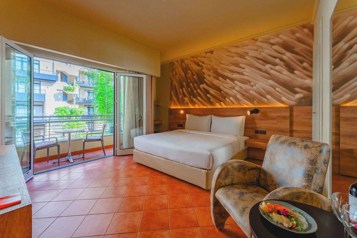 Una habitació d'hotel moderna amb un llit, un sofà, una taula de menjador amb un plat de menjar i un balcó que ofereix vistes a la vegetació i els edificis.