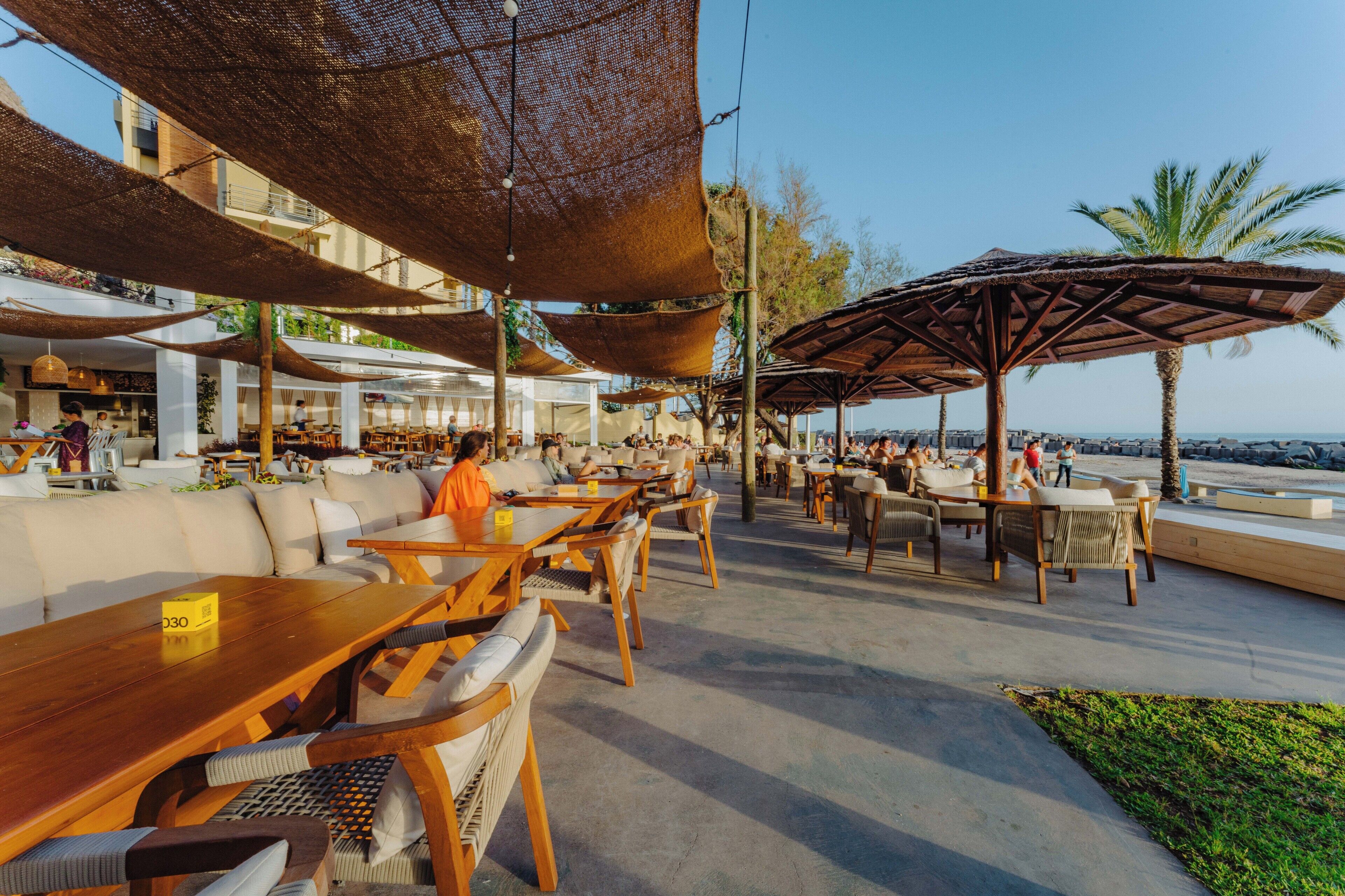 An outdoor dining area with wooden tables and chairs, shaded by large umbrellas, overlooking a body of water.