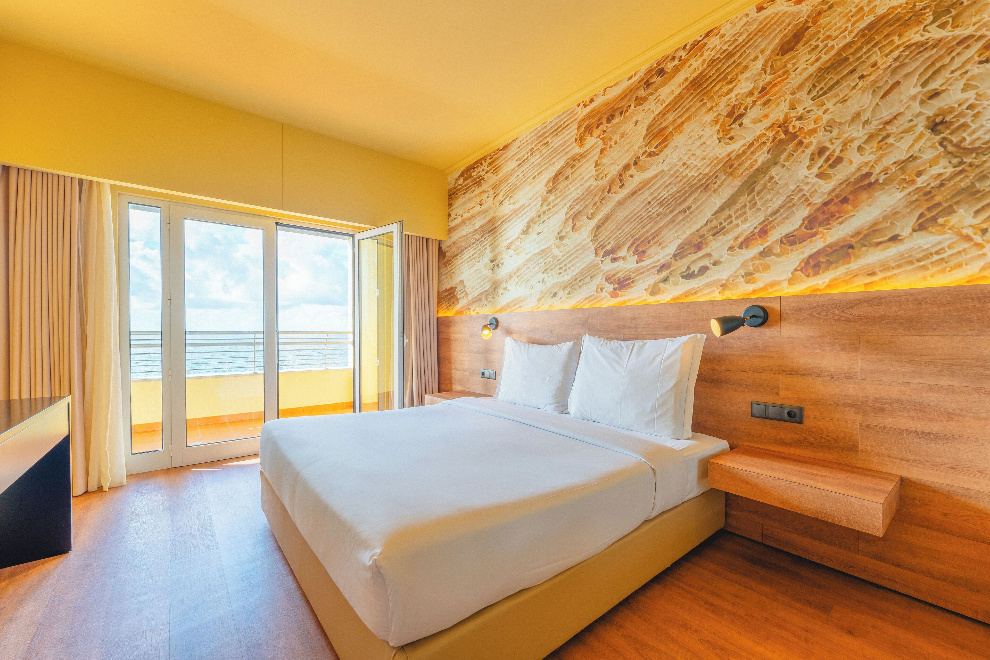 Habitació d'hotel moderna amb un llit gran, amb fusta al capçal i vista al mar a través de les portes corredisses de vidre.