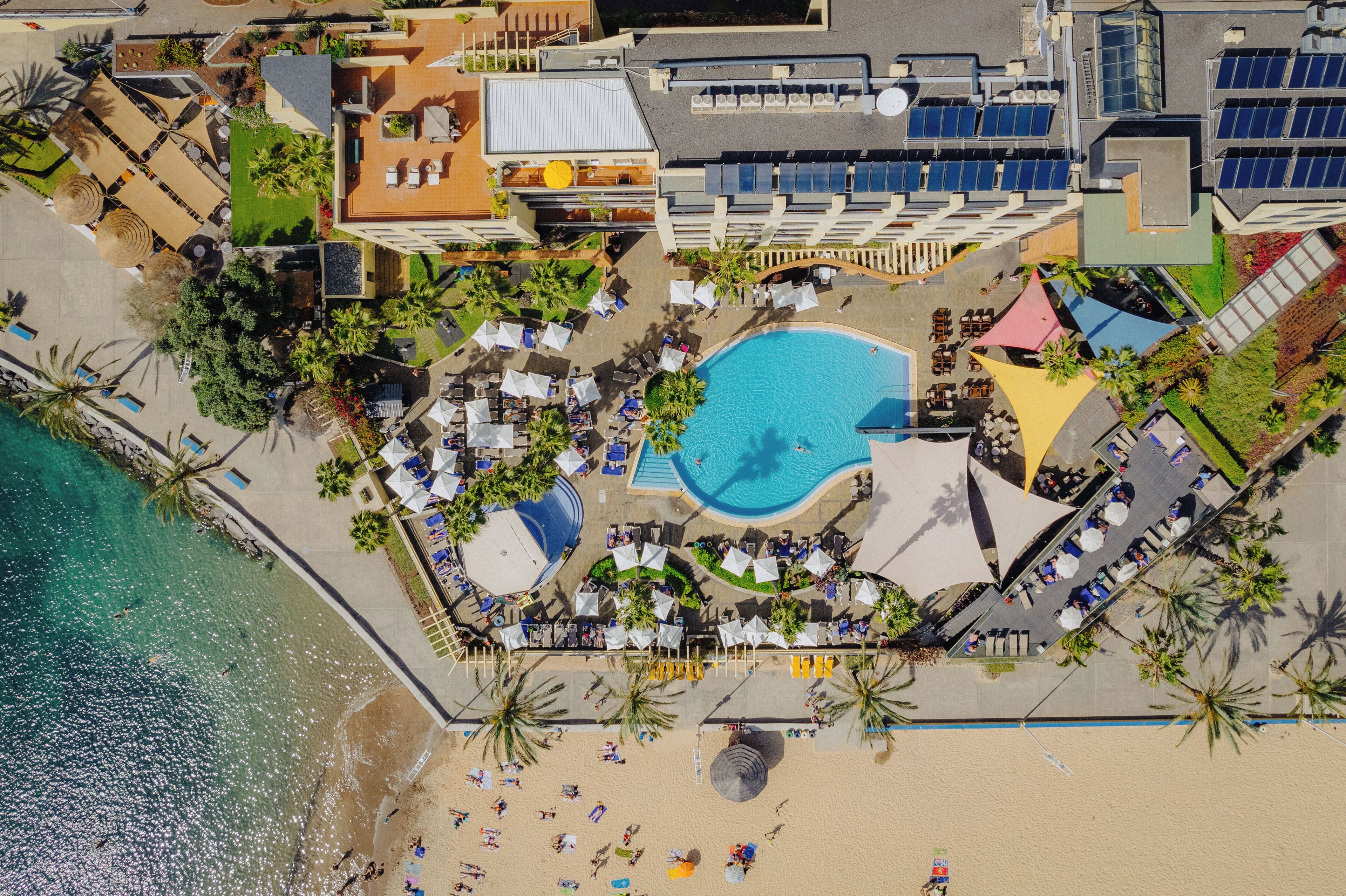 An aerial view of a resort with a swimming pool surrounded by lounge chairs and umbrellas, a beach area with sun loungers, and a building with solar panels.