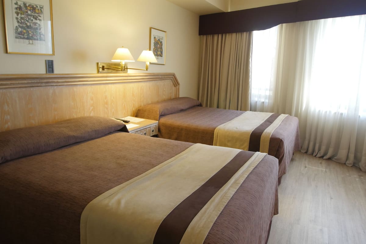Una habitación de hotel con dos camas, una mesita de noche, una lámpara y un ventanal con cortinas.