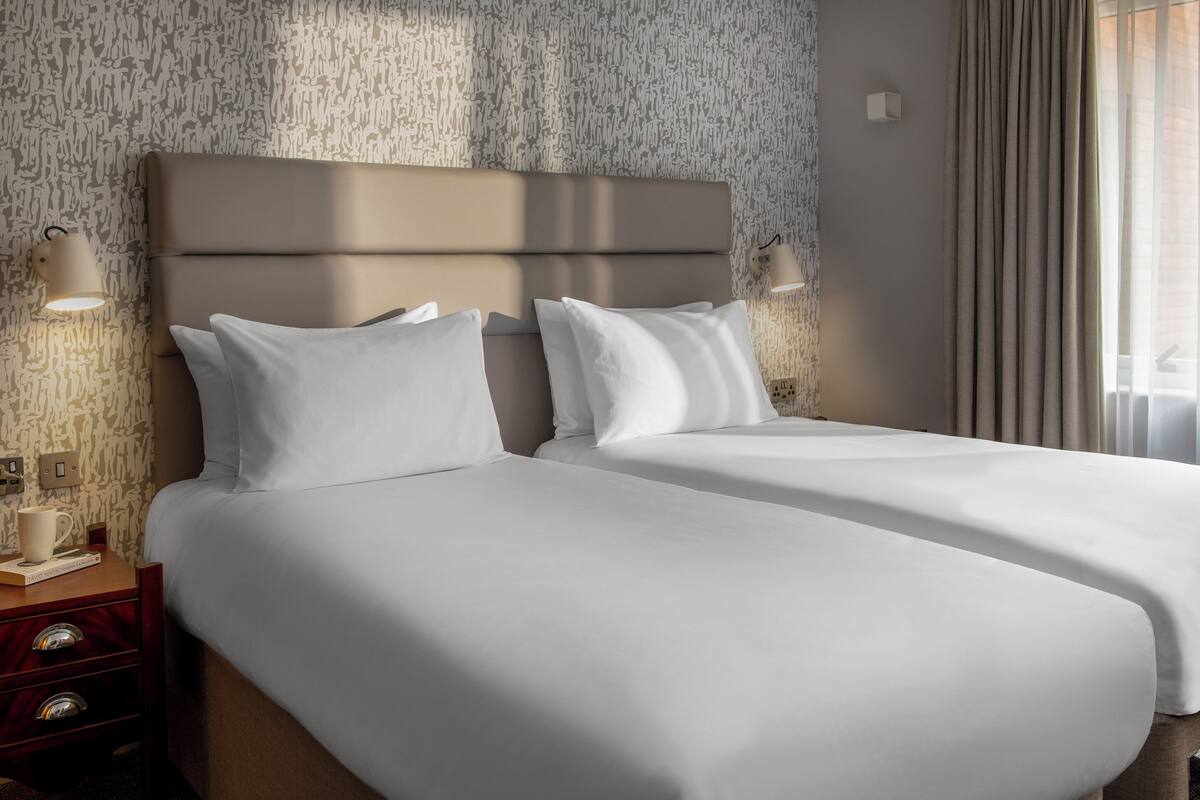 A hotel room with two beds, a nightstand with a lamp, a cup, and a book.
