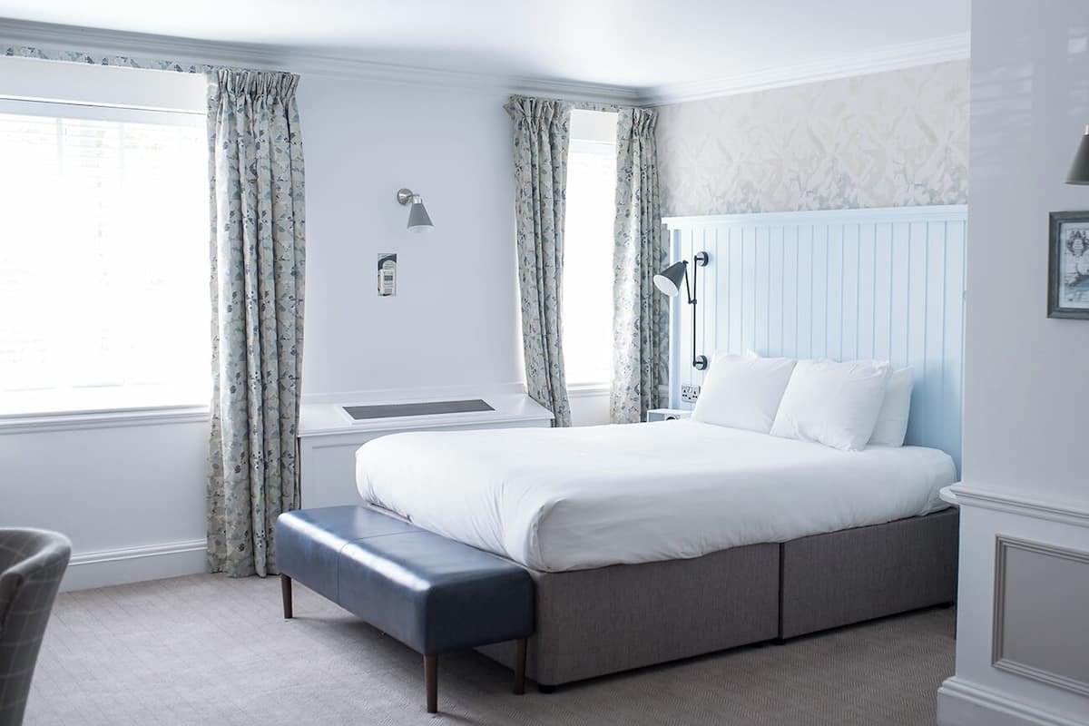 A bedroom with a bed, bench, bedside table, lamp, and curtains.