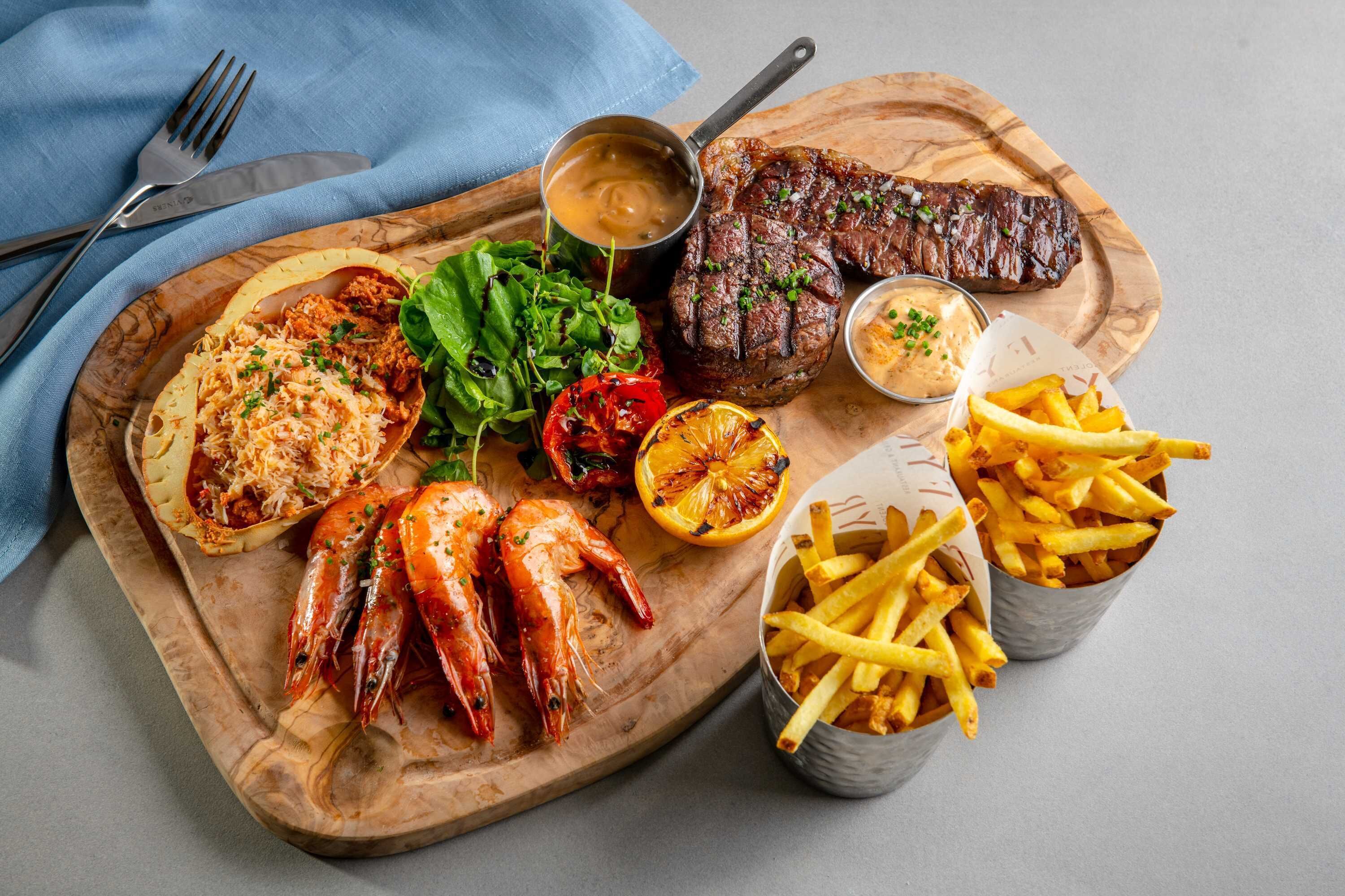 Una tabla de madera con bistec a la parrilla, camarones, ensalada, verduras asadas y papas fritas.