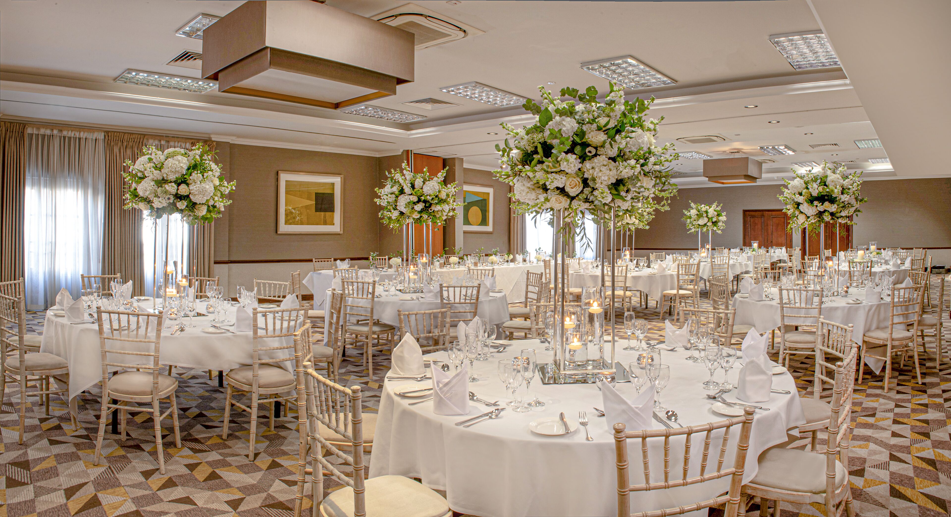 Un salón de banquetes con mesas redondas, manteles blancos y centros de mesa florales.