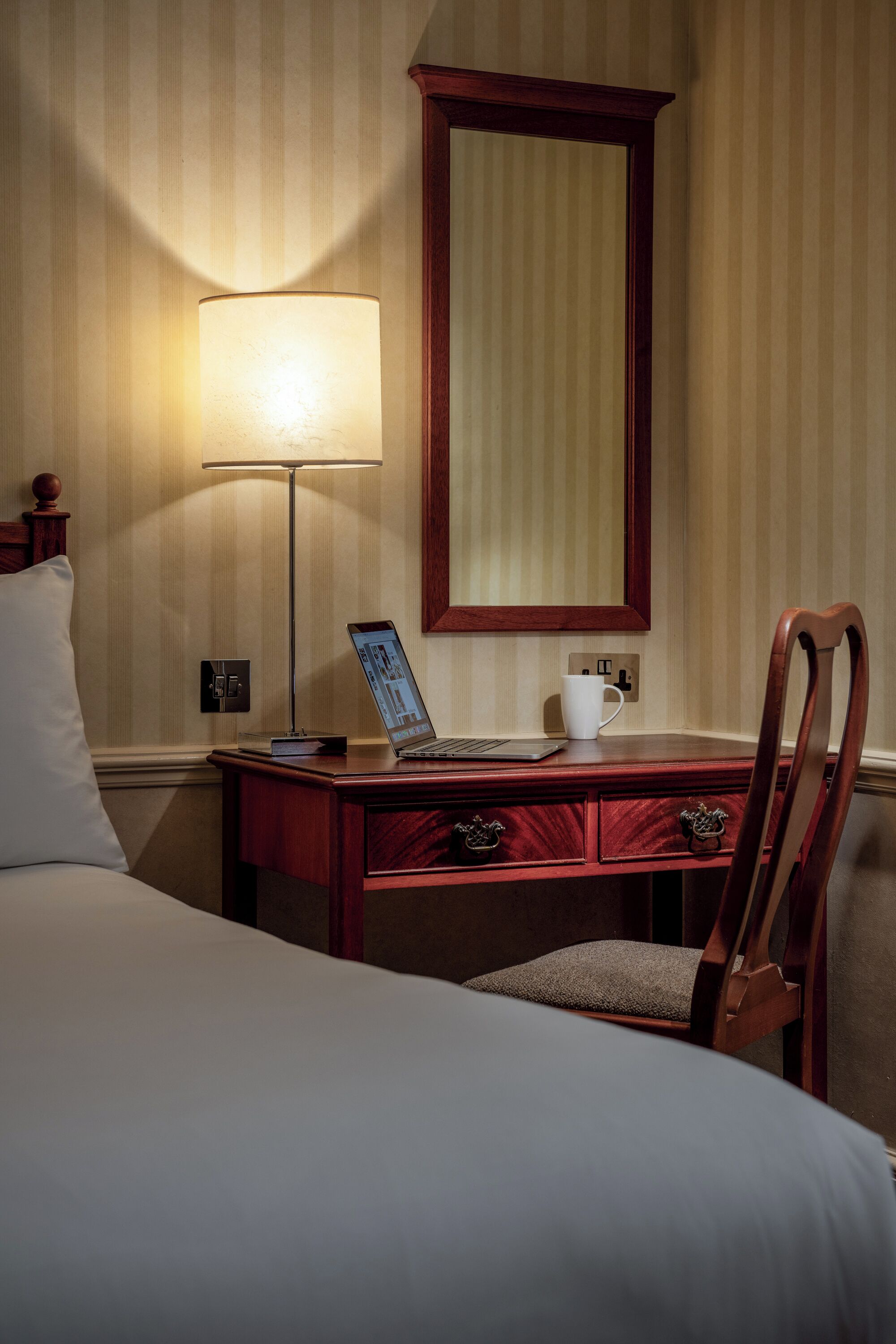 Quarto de hotel com uma cama, uma escrivaninha com um laptop, uma lâmpada e um espelho.