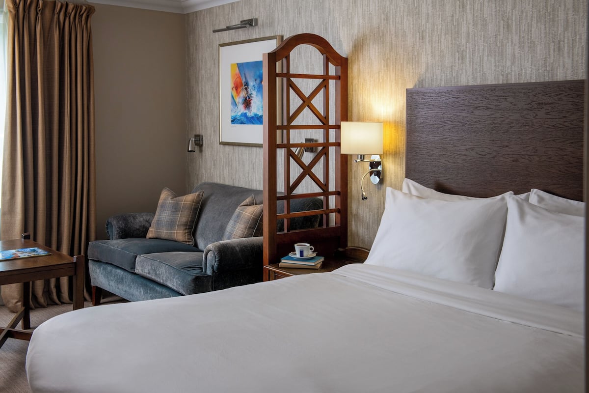 A hotel room with a large bed, a sofa, a wooden screen, a wall-mounted lamp, and a framed artwork.