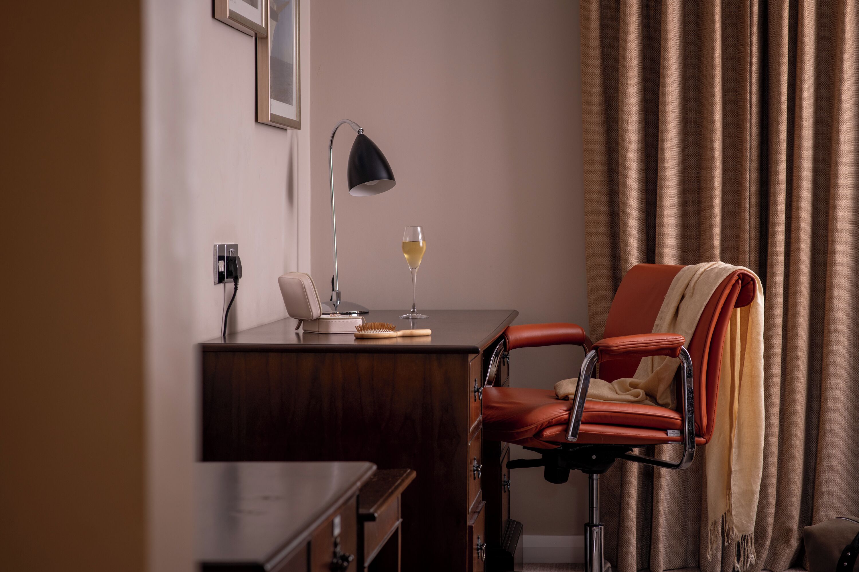 Habitación de hotel con escritorio, una silla, lámpara, una copa de vino y ventana con cortinas.
