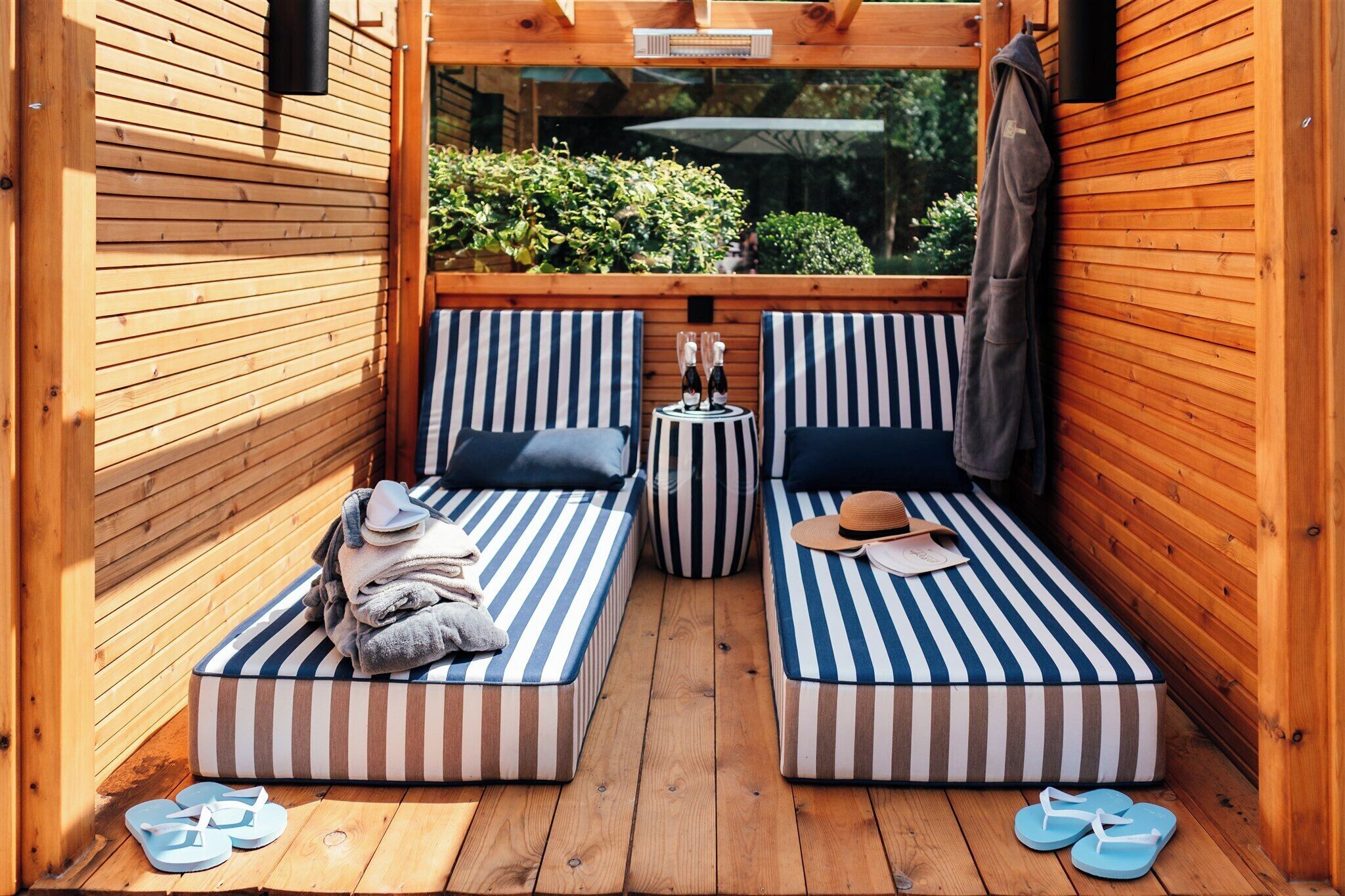 A wooden deck with two striped lounge beds, a small table with bottles, and a pair of flip-flops on the floor.