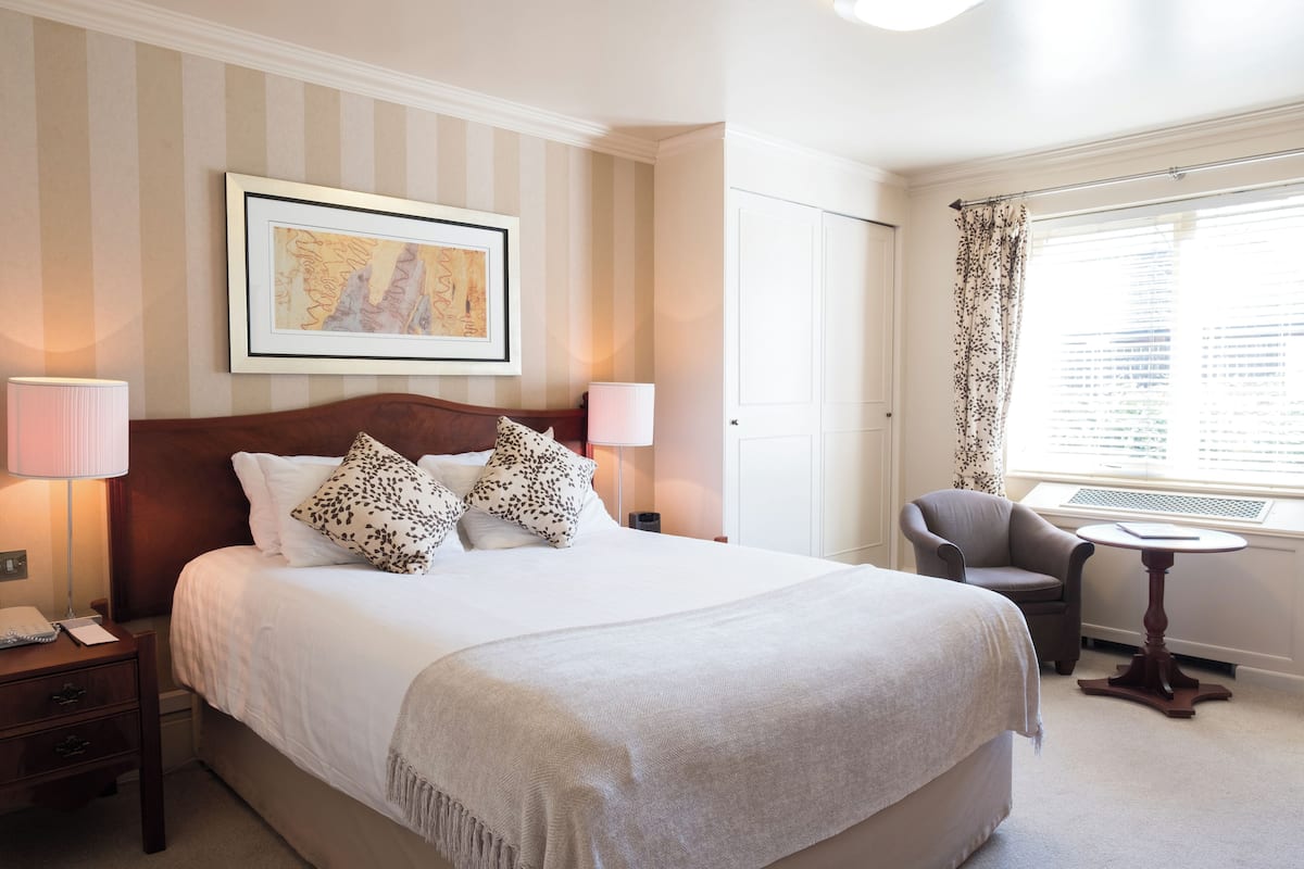 A hotel room with a large bed, two bedside lamps, a chair, a small table, a framed picture on the wall, and a window with curtains.