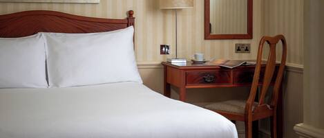 Een net opgemaakt bed met witte lakens, een houten nachtkastje met een lamp, een stoel en een ingelijst beeld op de muur.
