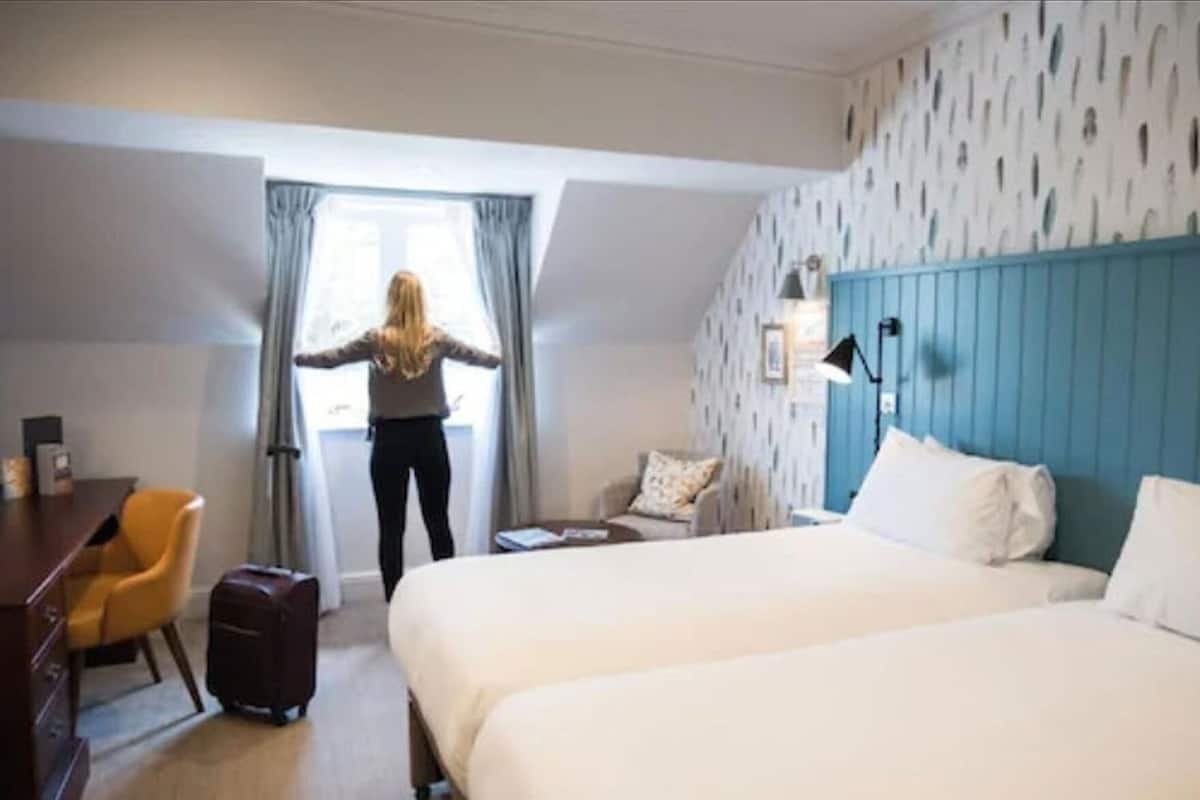 A hotel room with two beds, a desk, a chair, a suitcase, and a window with curtains.