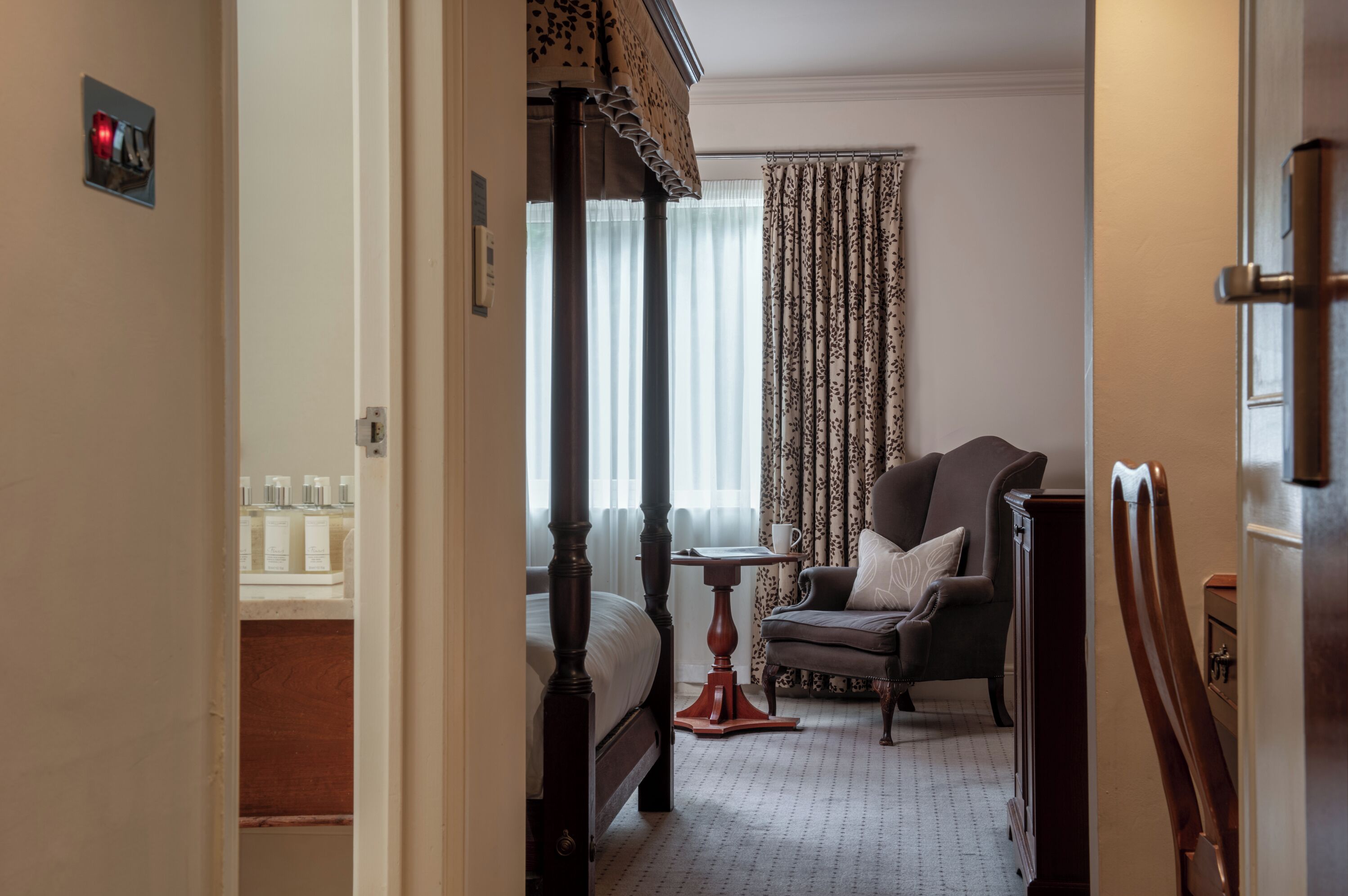 Une chambre d’hôtel avec un lit à baldaquin, un coin salon avec un fauteuil et une table, et une grande fenêtre avec des rideaux.