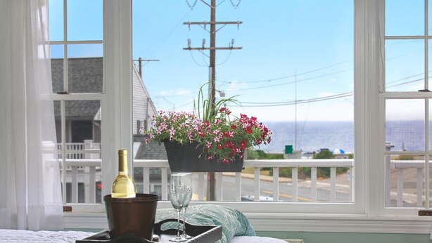 A bedroom with a bed, a window with a view of the ocean, a flower pot, a bottle of wine, and a glass on a tray. - Ocean Inn on Nantasket Beach (Hull)