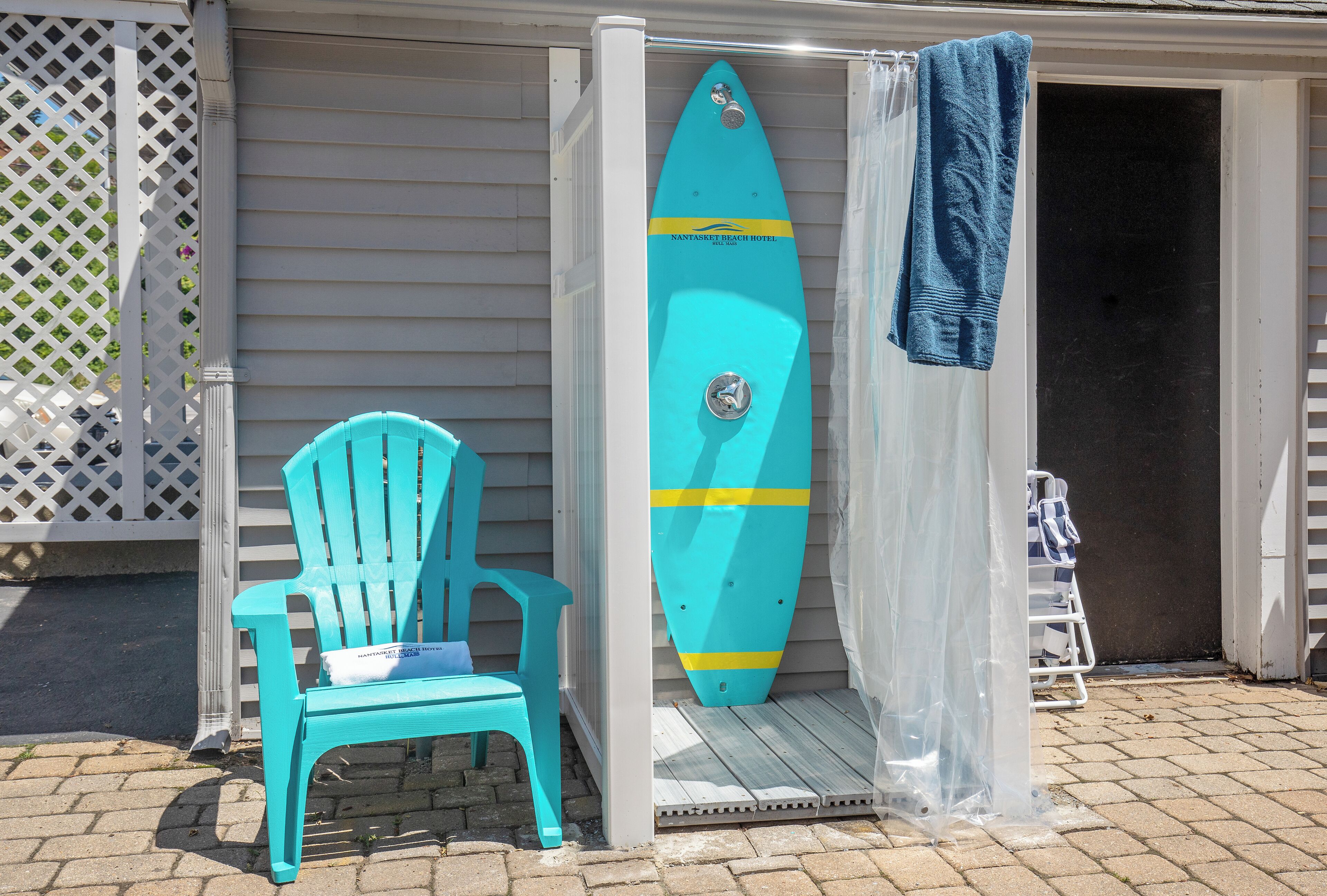 Una tavola da surf color turchese appoggiata a un muro bianco, una sedia blu e una tenda da doccia.