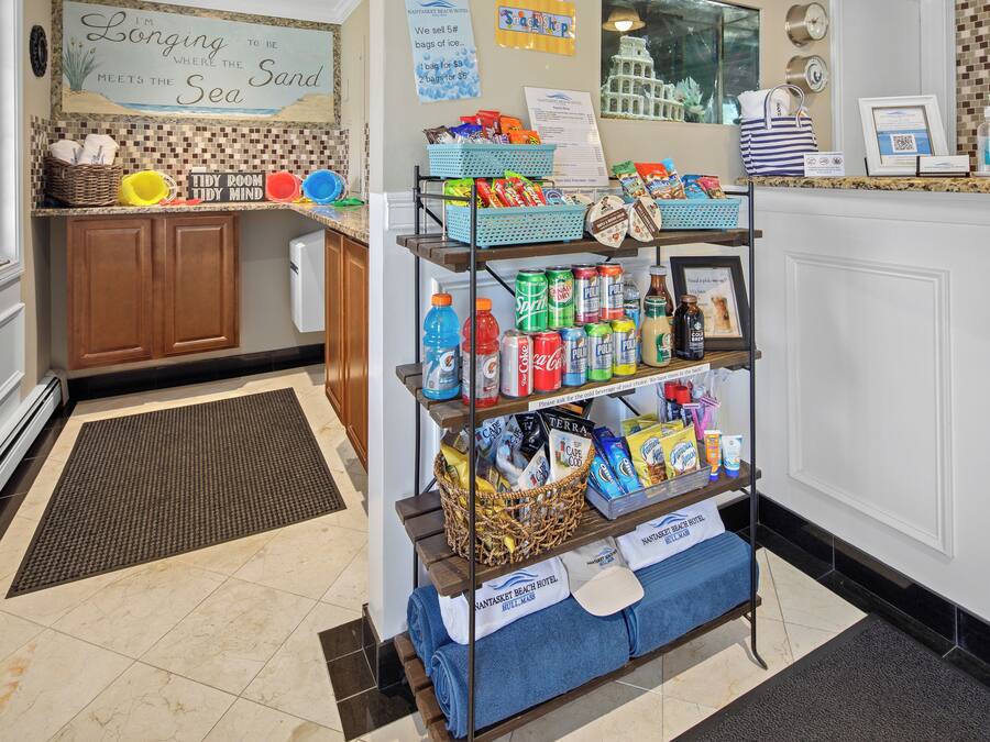 A hotel reception area with a self-service kiosk stocked with snacks and beverages, a sign reading "I'm Longing to be Where the Sea Meets the Sand," and a tiled floor.