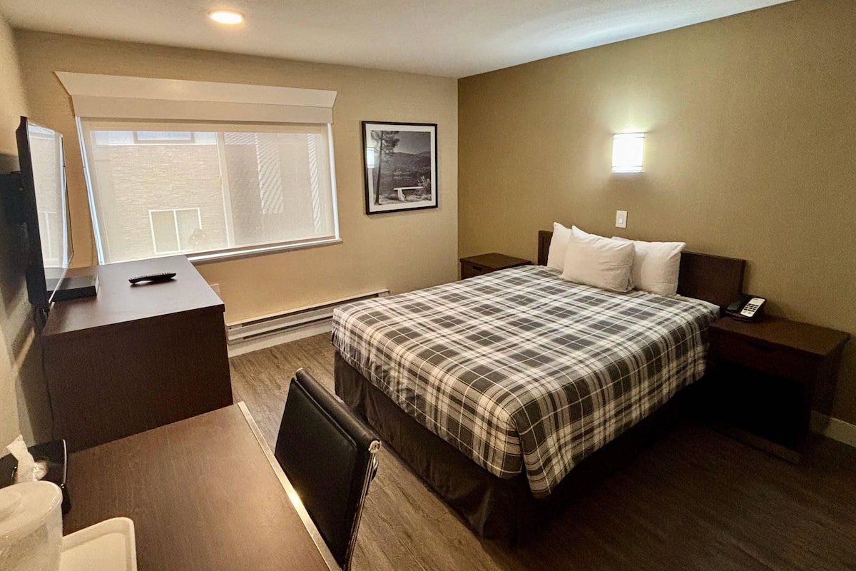 A hotel room with a bed, bedside table, desk, chair, and a window with blinds.