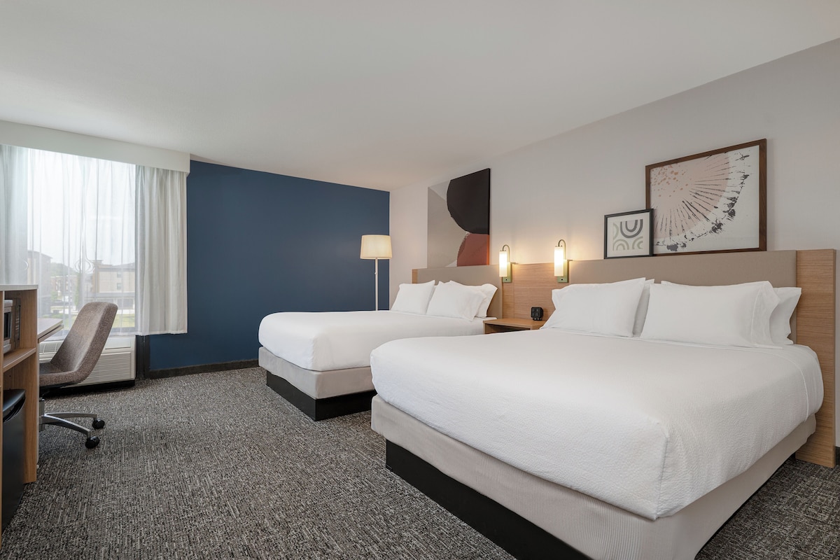 Quarto de hotel com duas camas, uma escrivaninha, uma cadeira e uma janela grande com cortinas.