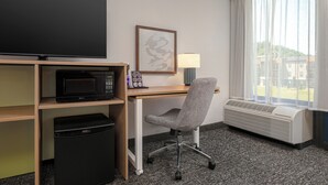 A hotel room with a desk, television, microwave, and air conditioning unit.