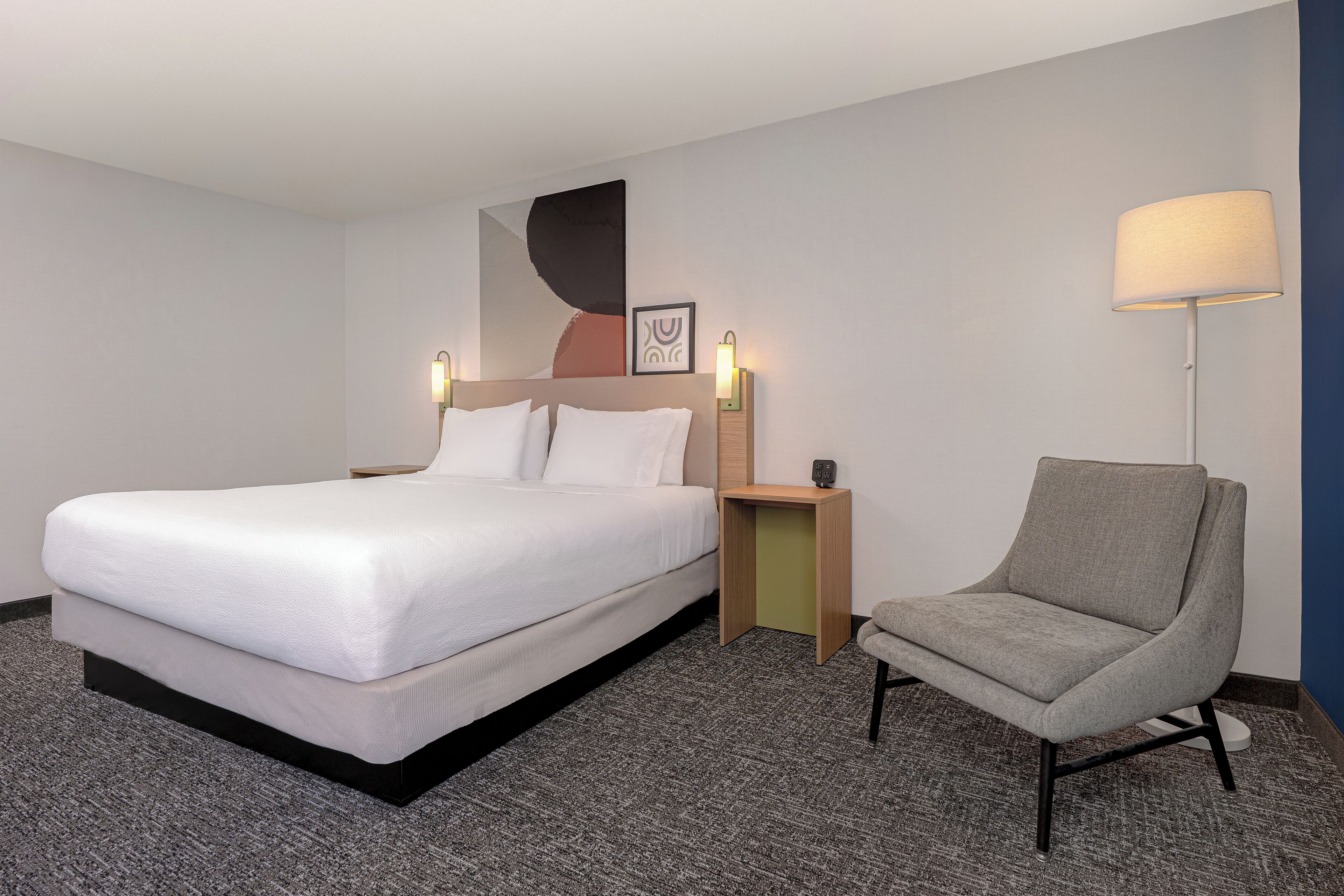 A hotel room with a bed, a chair, a nightstand, a wall art piece, and a lamp.