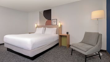 A hotel room with a bed, a chair, a nightstand, a wall art piece, and a lamp.
