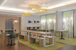 Uma área de jantar moderna com várias mesas e cadeiras, teto com luminárias pendentes e vista para o exterior através de grandes janelas.