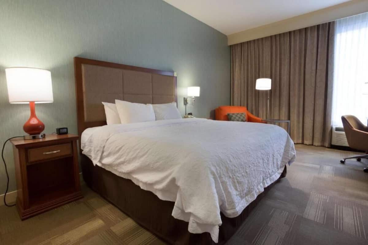A hotel room with a large bed, a nightstand with a lamp, a chair, and a window with curtains.