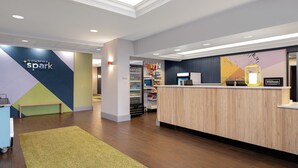 Et hotellets receptionområde med en trædisk, et siddeområde med en bænk, og en væg med et geometrisk design samt et skilt, der siger 'It only takes a spark'.