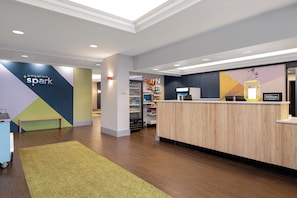 Uma área de recepção de hotel com um balcão de madeira, um espaço de estar com um banco e uma parede com design geométrico e um letreiro que diz 'It only takes a spark'.