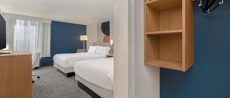 Ett hotellrum med två sängar, ett skrivbord, en stol, en klädställning och en hylla som är monterad på väggen.