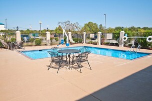 A swimming pool with lounge chairs and a table, surrounded by a fence and greenery.