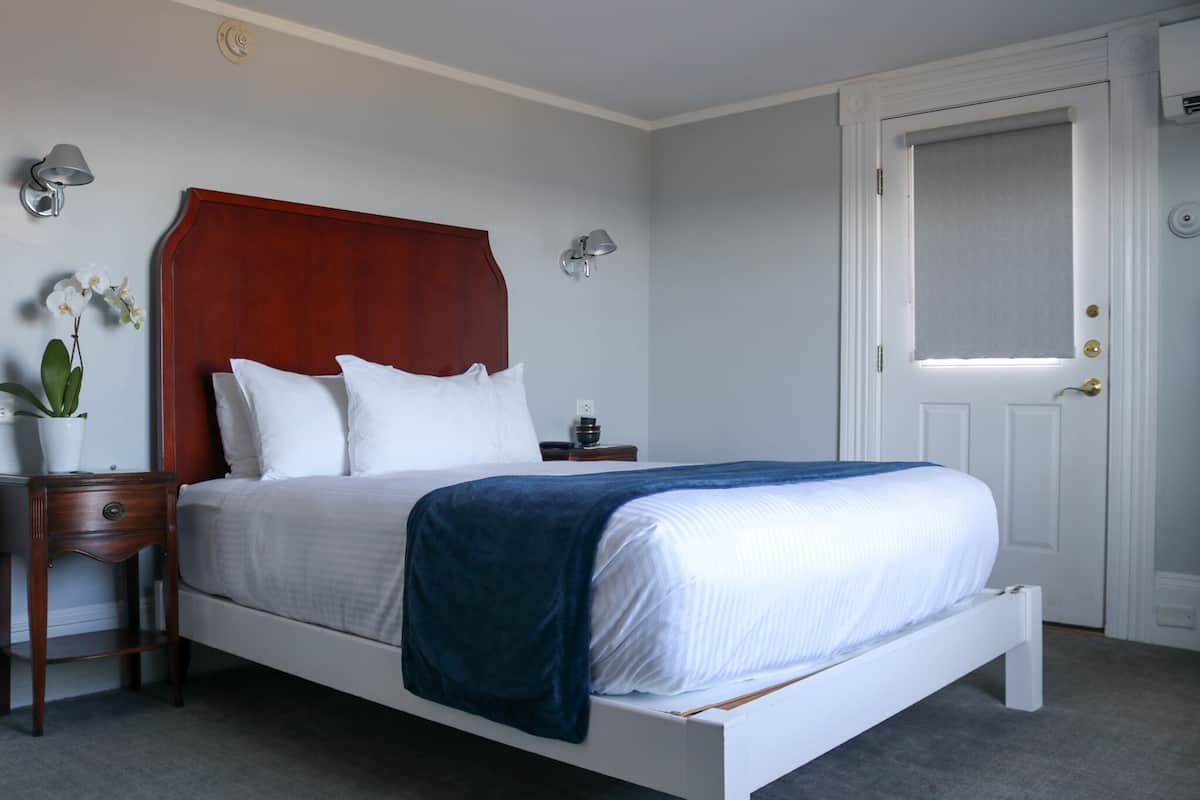 A bedroom with a white bed, a wooden headboard, a nightstand with a lamp, and a door with a window.