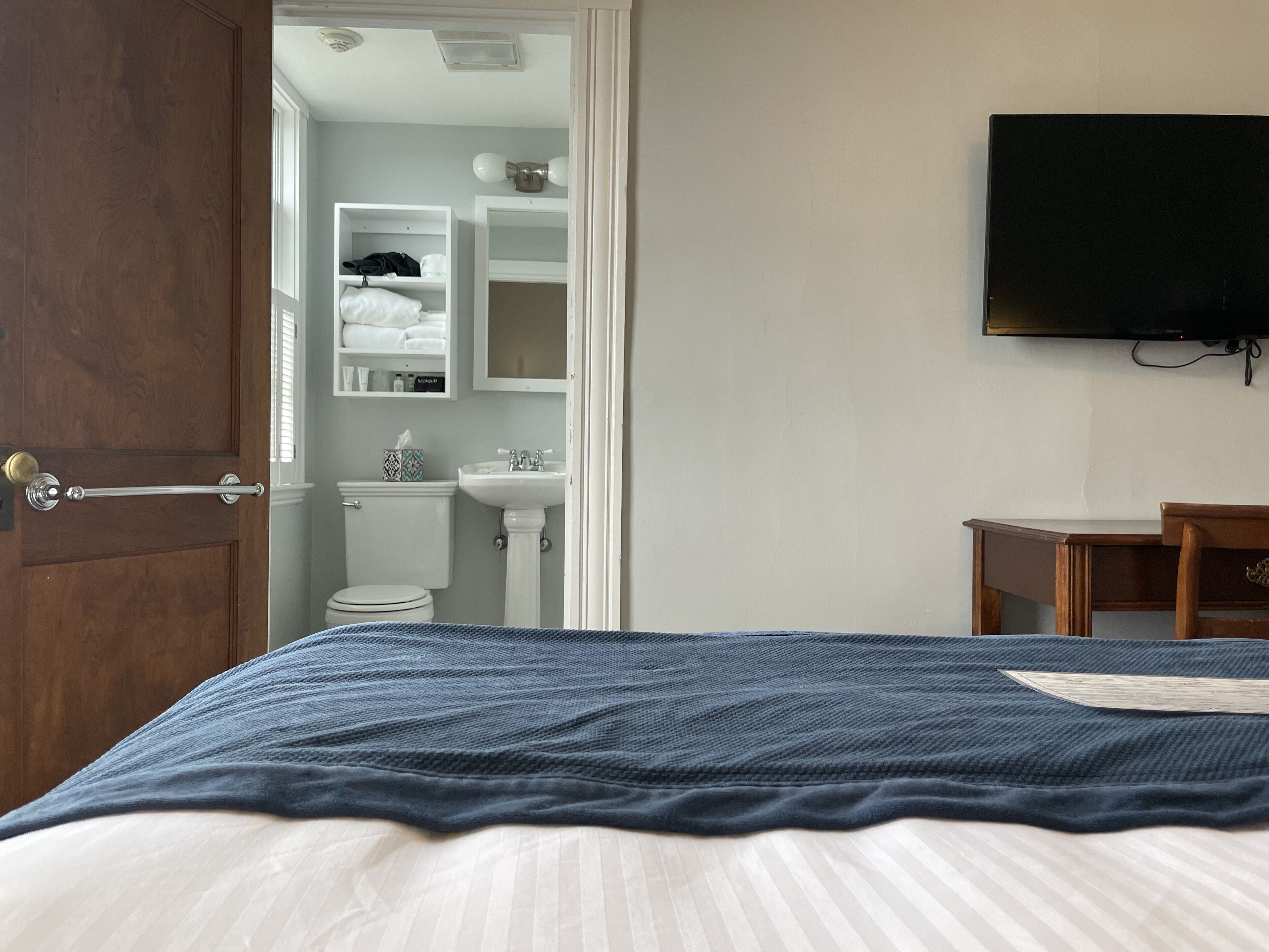 Un dormitorio con cama, una mesita de noche de madera, televisión y un baño visible a través de una puerta abierta, además de un lavabo blanco.