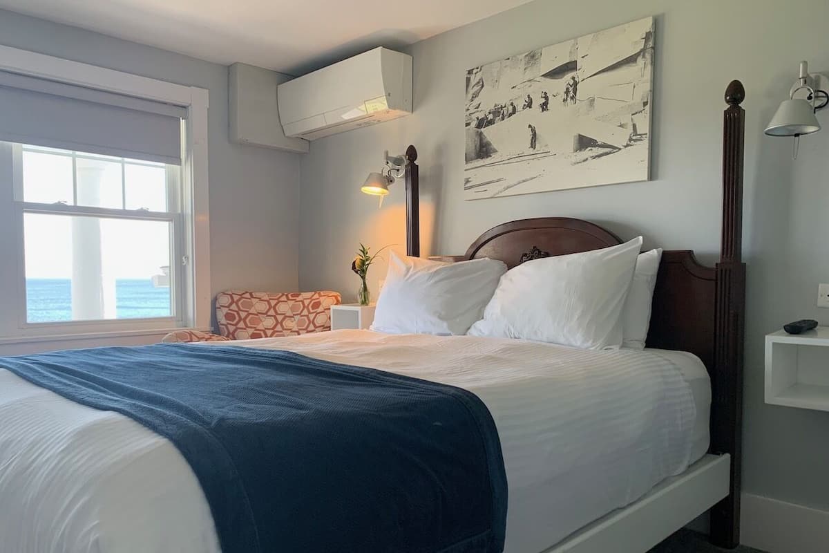 A neatly made bed with a blue blanket, a wooden headboard, and a wall-mounted air conditioner.