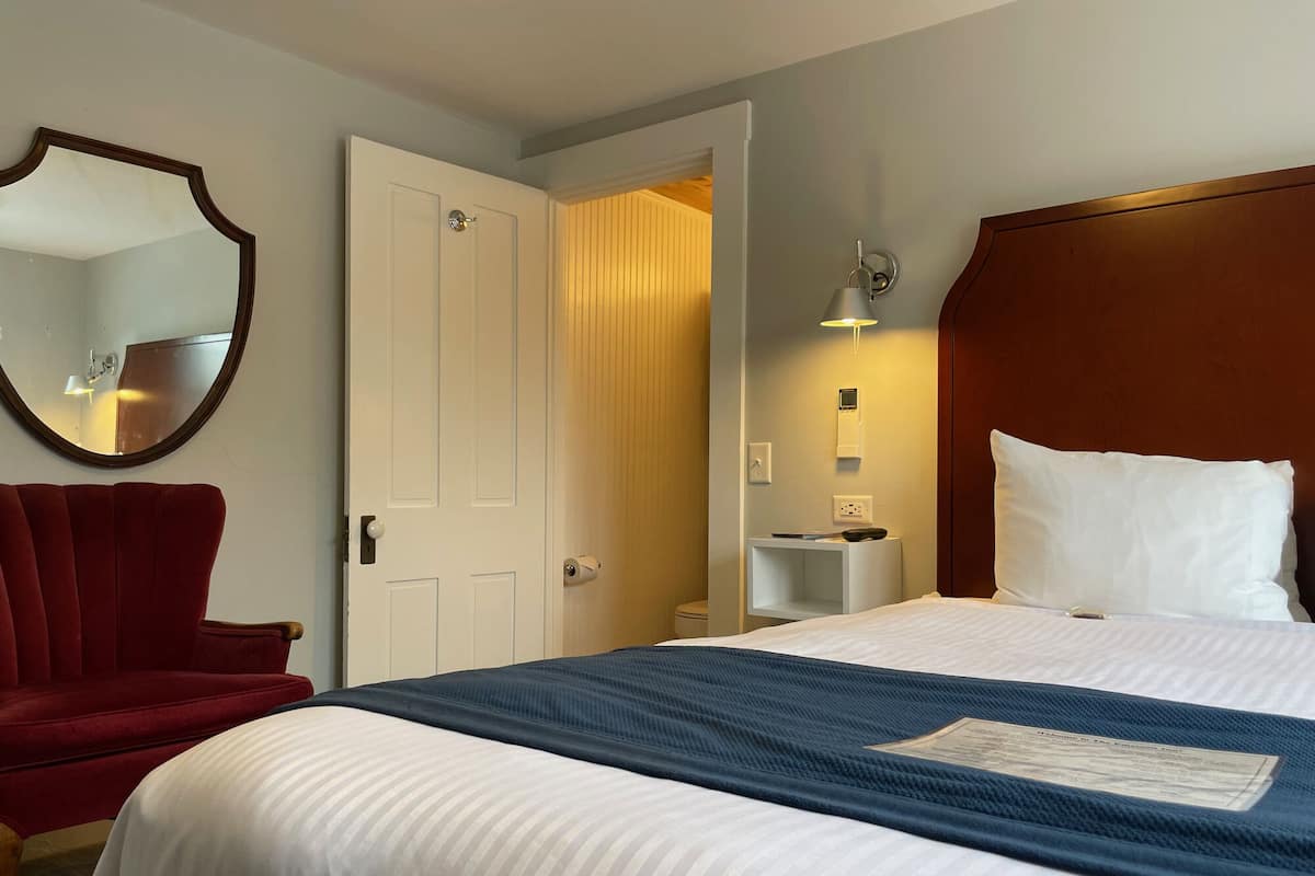 A hotel room with a bed, a red chair, a white door, and a wall-mounted mirror.
