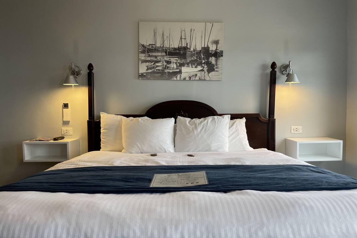 A neatly made bed with white and blue bedding, two bedside tables with lamps, and a black and white maritime painting on the wall.