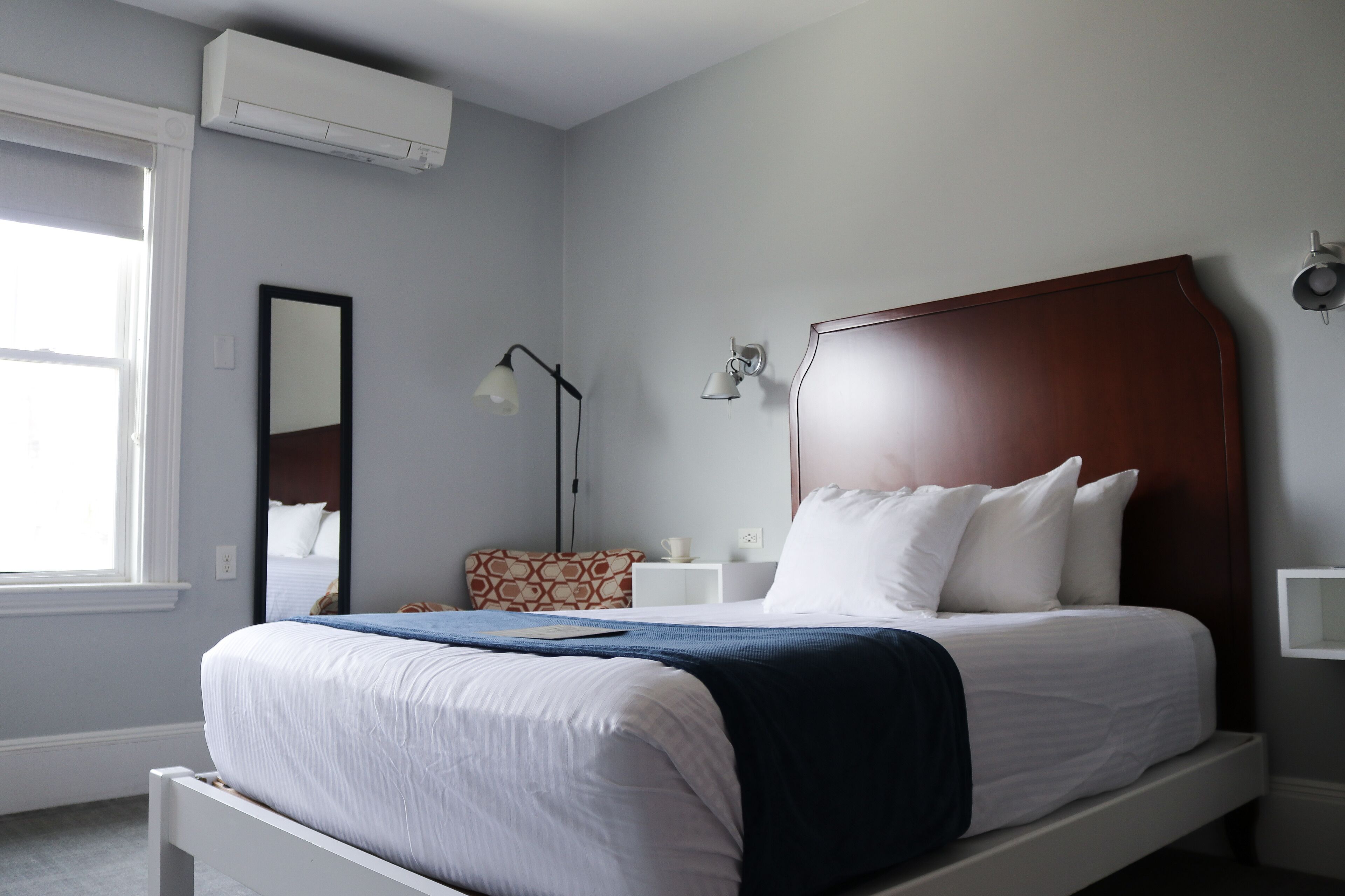 A neatly made bed with white bedding and a blue throw, a bedside table with a lamp, a wall-mounted air conditioner, and a window with a white frame.