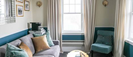 Ein Wohnzimmer mit einer Couch, einem Couchtisch und einem Sessel. Vorhanden sind Vorhänge, ein Fenster und Wanddekorationen.