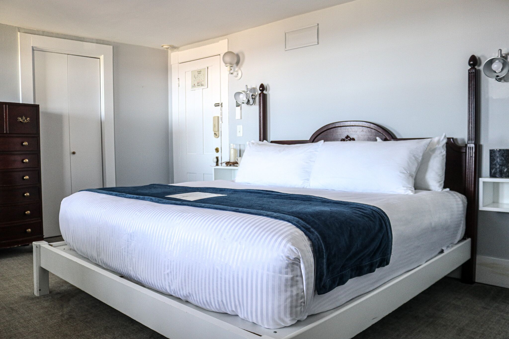 A single-bed room with a wooden headboard, a white mattress, and a blue blanket. There is a wooden dresser to the left and a nightstand with a lamp to the right.
