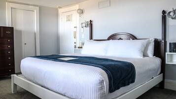 A single-bed room with a wooden headboard, a white mattress, and a blue blanket. There is a wooden dresser to the left and a nightstand with a lamp to the right.