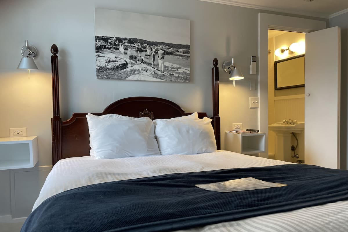 A neatly made bed with white linens and a dark blue throw blanket, a bedside table with a lamp, a wall-mounted picture, and a bathroom visible through an open door.
